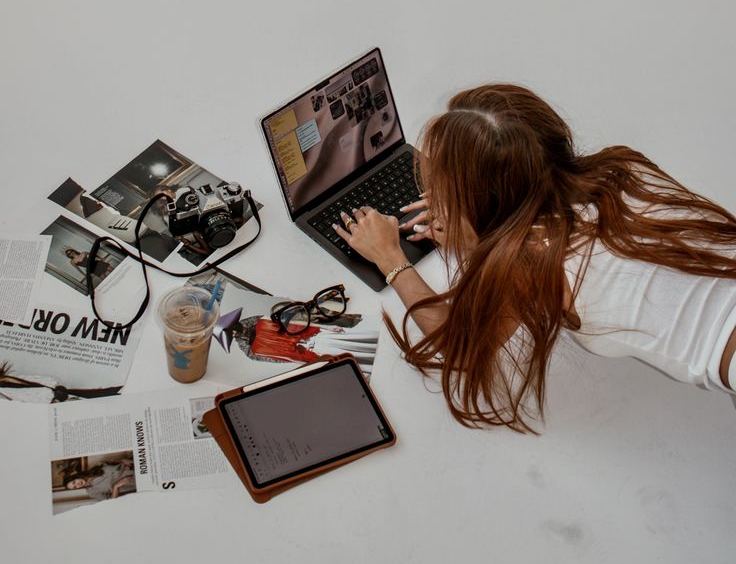 A content creator at a laptop surrounded by prints, a camera and a tablet